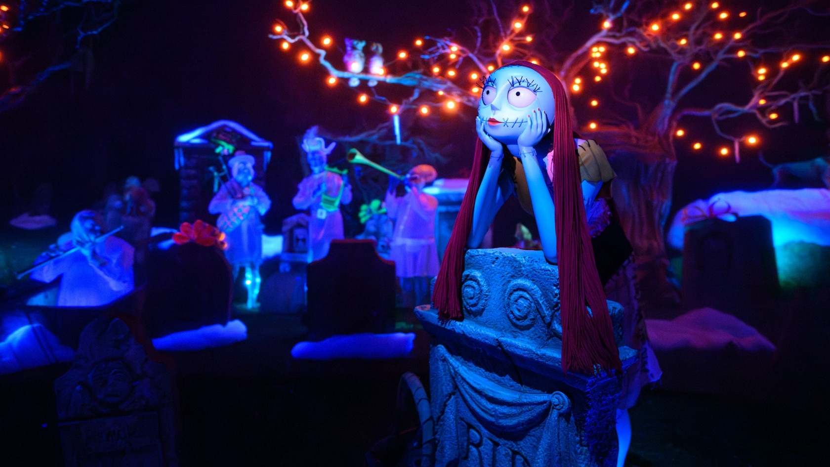 Sally from The Nightmare Before Christmas leaning on a stone pedestal in the Haunted Mansion Holiday graveyard, with glowing ghost figures and a tree strung with orange lights behind her