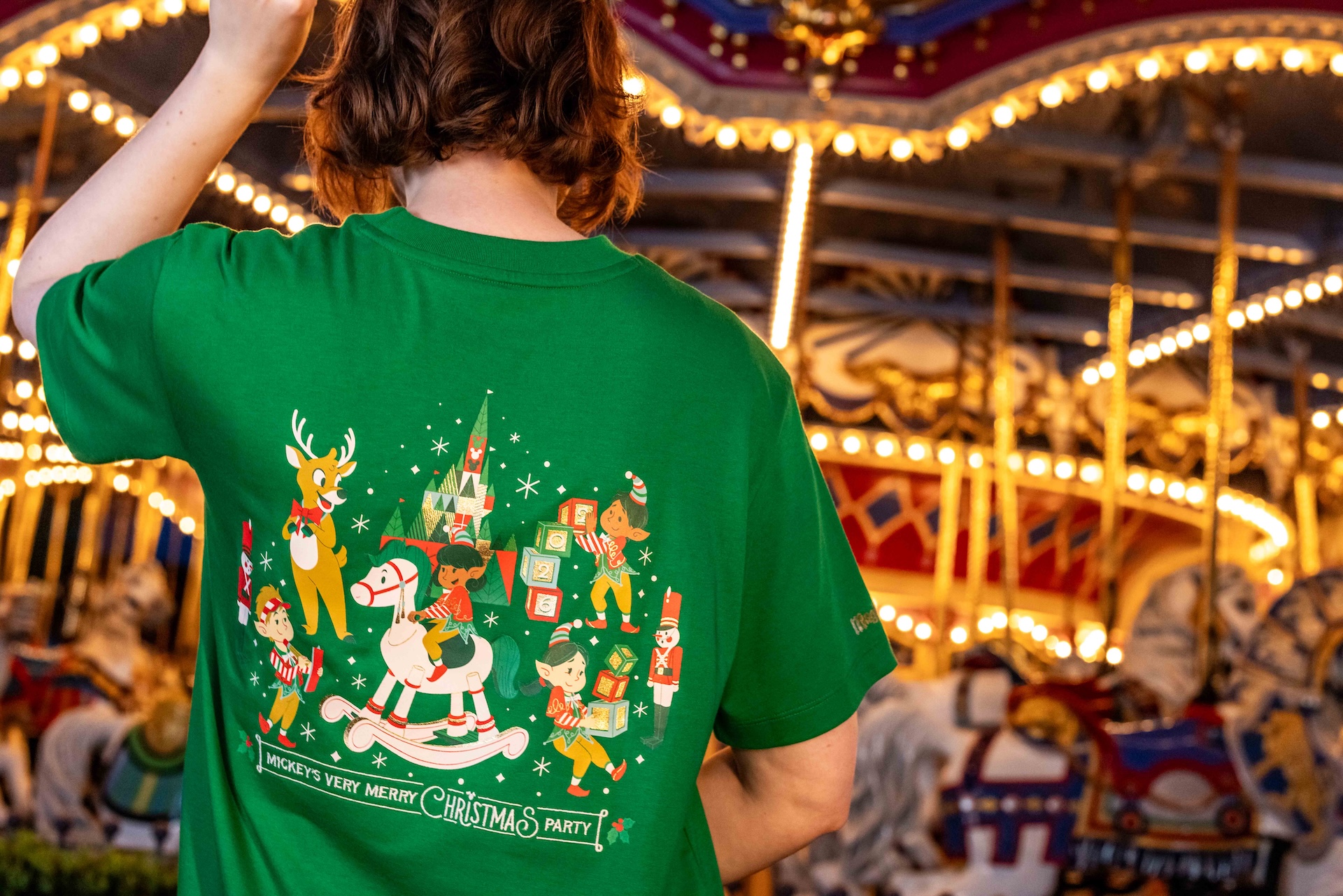 Back of a green 2026 Mickey's Very Merry Christmas Party tee showing elves, a reindeer, a rocking horse, toy soldiers and blocks spelling 2026 in front of a castle