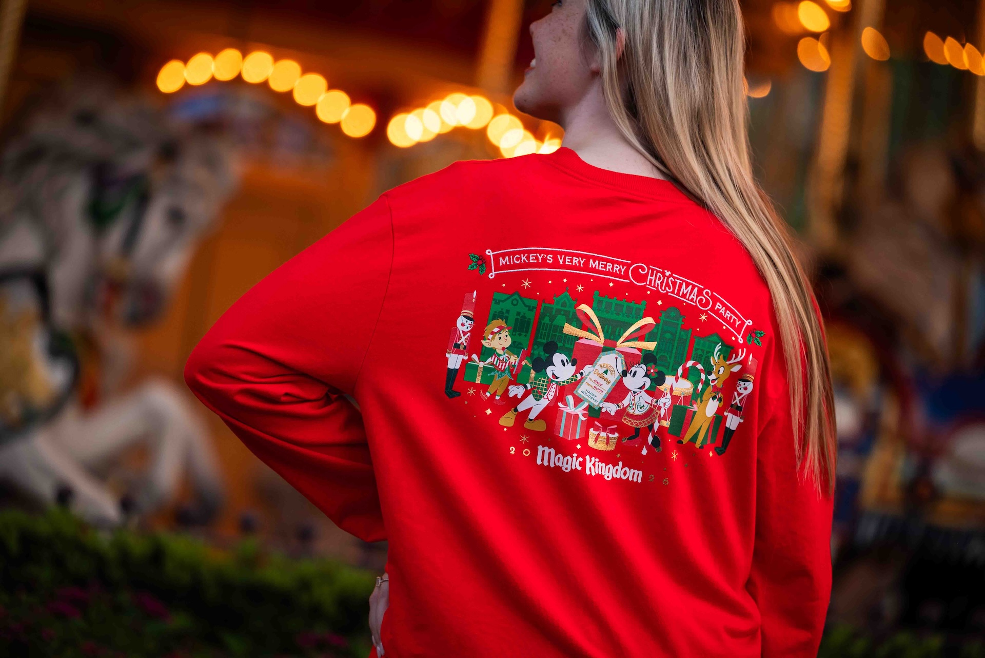 Back of the red 2026 Mickey's Very Merry Christmas Party pullover showing Mickey Mouse, Minnie Mouse, an elf, a reindeer and two toy soldiers with a large gift, above the words Magic Kingdom