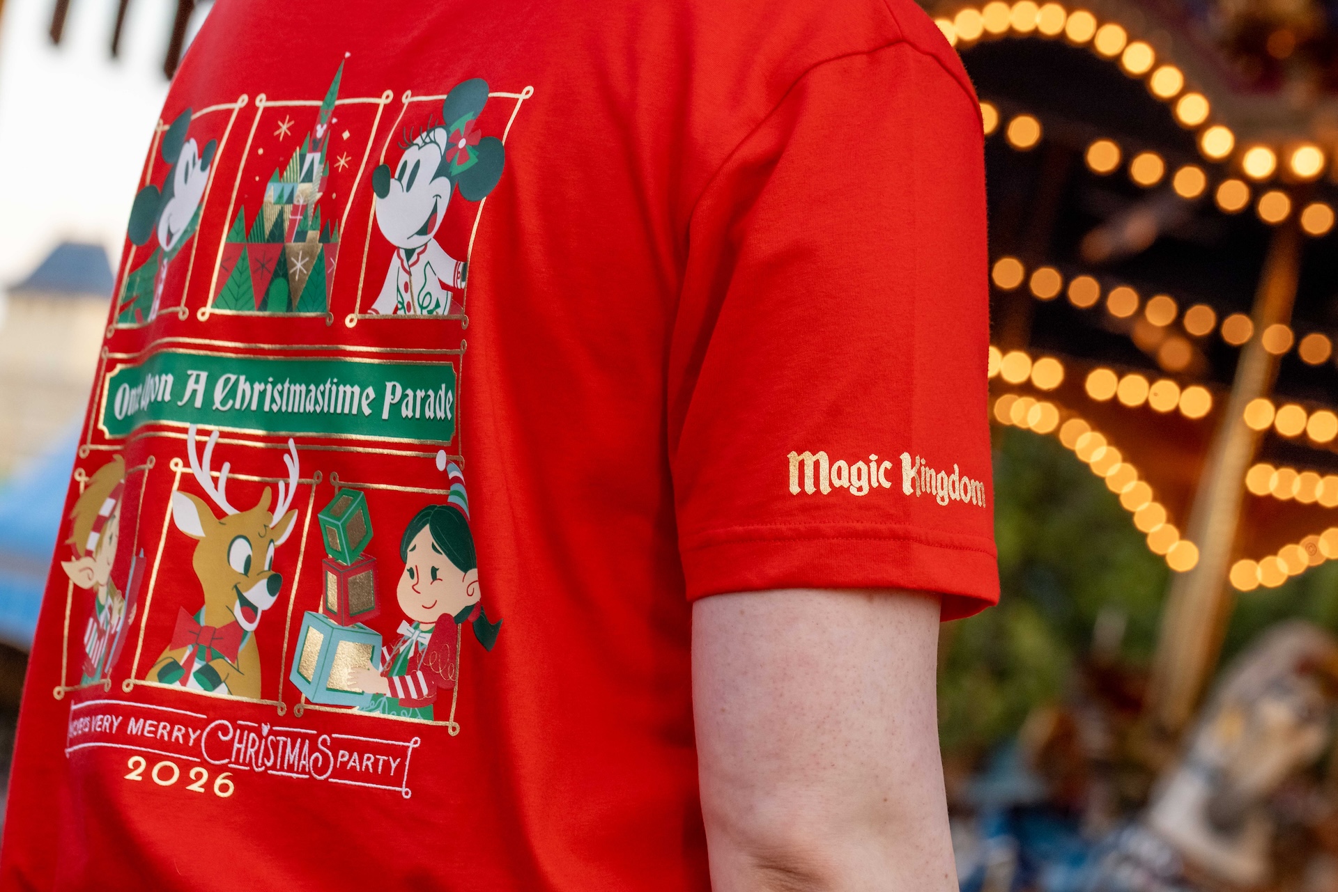 Red 2026 Mickey's Very Merry Christmas Party tee with framed art of Mickey Mouse, Minnie Mouse, a castle, a reindeer and elves, a Once Upon A Christmastime Parade banner, and Magic Kingdom printed in gold on the sleeve