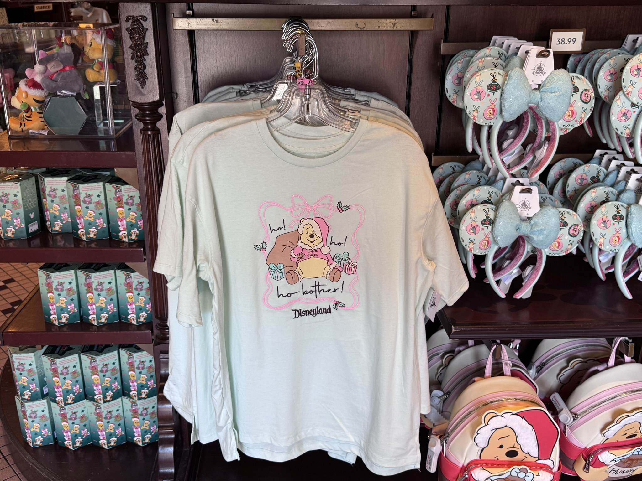 Mint green Disneyland t-shirt showing Winnie the Pooh in a pink Santa suit with the words ho ho ho bother, beside Pooh ornament Minnie ears and Santa Pooh backpacks at the Emporium