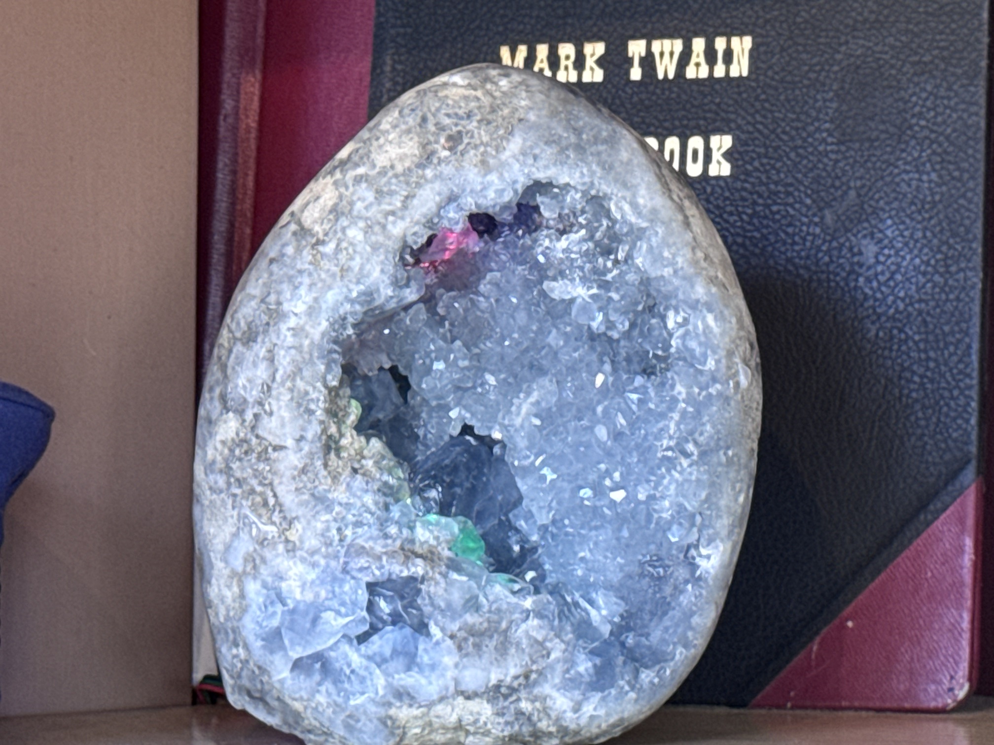 Pale blue crystal geode with a lit cavity on a shelf beside a Mark Twain book, a brass bell, a conductor's cap and a Samuel Clemens pilot's certificate