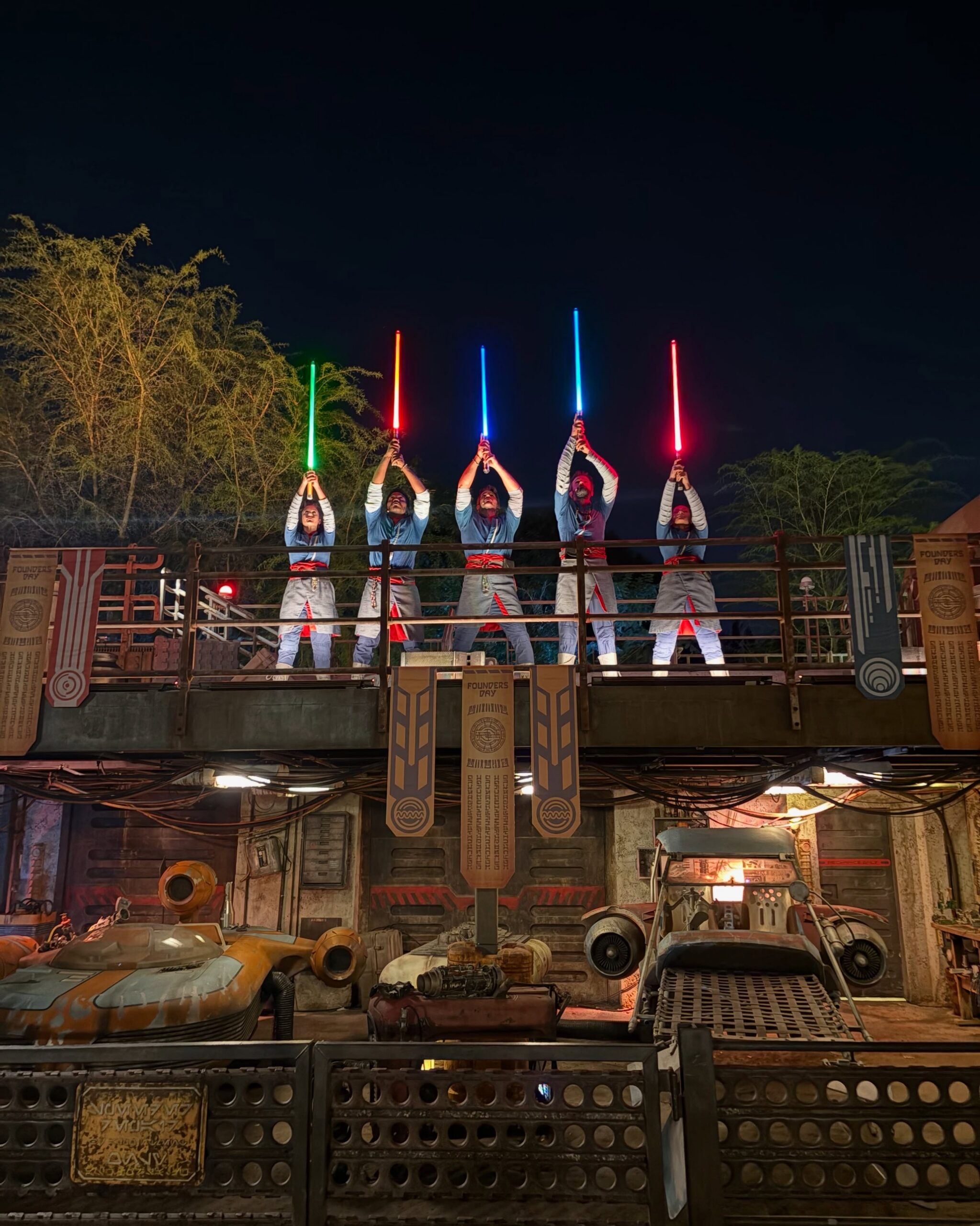 Five Saja raise lit lightsabers overhead on a platform draped with Founders Day banners in Star Wars: Galaxy's Edge at Disney's Hollywood Studios
