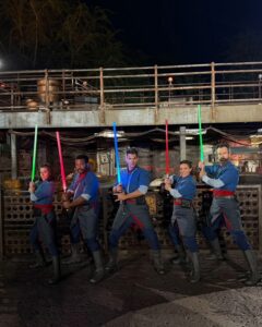 Five Saja in blue and gray robes with red sashes pose with lit lightsabers at night in Star Wars: Galaxy's Edge at Disney's Hollywood Studios